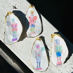 May include: Four oyster shells with gold trim, each painted with a different nutcracker design. The nutcrackers are wearing colorful uniforms and hats, and holding various accessories like flowers and flags. The text "Merry Christmas" is written on one of the shells.