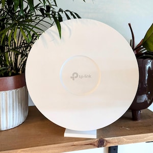 May include: White circular TP-Link Wi-Fi extender with the TP-Link logo on the front. The extender is sitting on a wooden shelf.