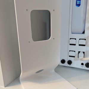 May include: A white, angled bracket with a rectangular cutout and a curved base. The bracket has a modern design and is positioned in front of a white electronic device with a screen displaying data.