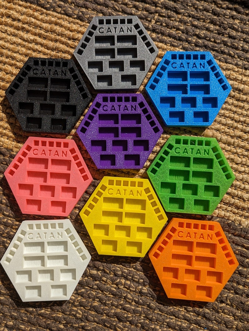 May include: Eight hexagonal Catan game tile organizers in different colors: gray, black, purple, pink, green, yellow, white, and orange. Each tile organizer has the word "CATAN" printed on it.