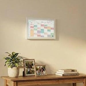 May include: A wooden console table with a framed calendar, two framed photos, a potted plant, and a stack of books. The calendar has a white frame and a colorful grid. The photos show family portraits.