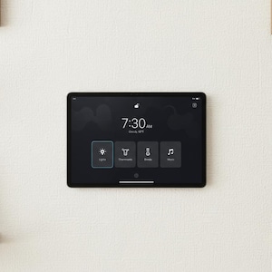 May include: A black tablet displaying a smart home interface, mounted on a white textured wall. The screen shows the time, weather, and control icons for lights, thermostat, blinds, and music. Wooden shelves and framed art are visible.