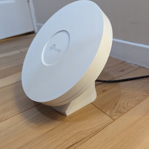 Może przedstawiać: Biały router bezprzewodowy TP-Link o okrągłym kształcie z białą podstawą. Router stoi na drewnianej podłodze.