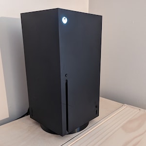 May include: A black gaming console with a white light on the front. The console is sitting on a wooden surface.