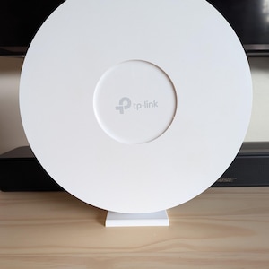 May include: A white TP-Link Wi-Fi access point. The device is circular with a smaller, recessed centre. The TP-Link logo is visible in the centre. It is mounted on a white stand and sits on a wooden surface.