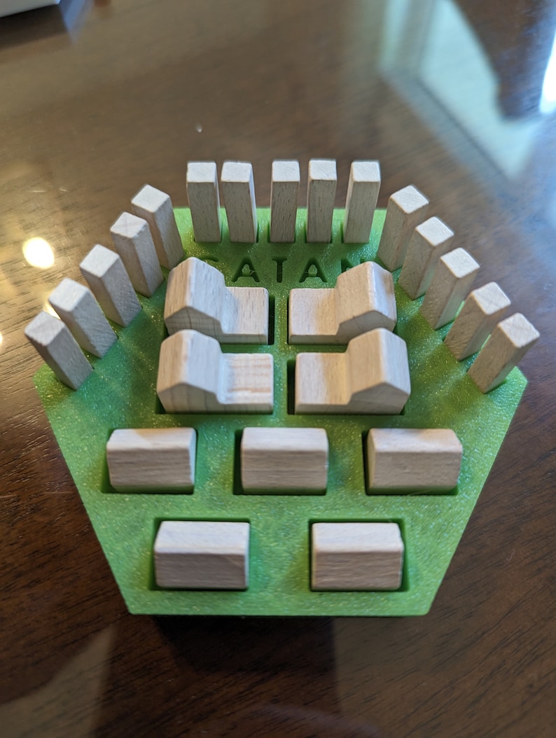 May include: A green hexagonal board game piece with the word "CATAN" printed on it. The piece has wooden houses and roads on it.