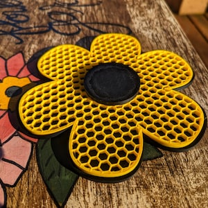 Puede incluir: Un posavasos amarillo en forma de flor con un patrón de panal. El posavasos tiene un centro negro.