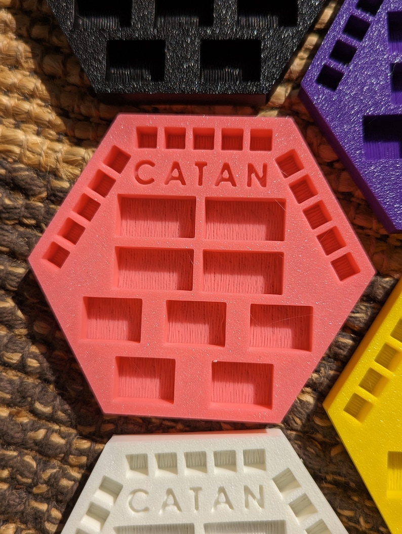 May include: A pink hexagonal Catan game tile organizer with the word "CATAN" printed on the top. The organizer has multiple compartments for storing game pieces.