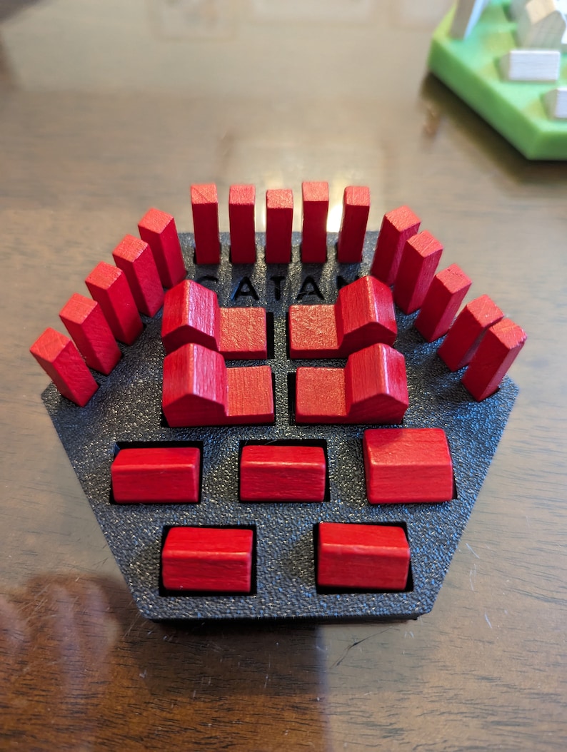 May include: A red wooden Catan game resource set with a black plastic base. The set includes 15 resource pieces, 6 houses, and 6 cities. The base has the word "Catan" printed on it.