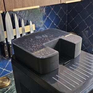Op de afbeelding: Zwarte espressomachine met een doorzichtige bonencontainer gevuld met koffiebonen. De machine heeft een strak design met een zilveren lekbak. Een set messen hangt op de achtergrond aan de muur.