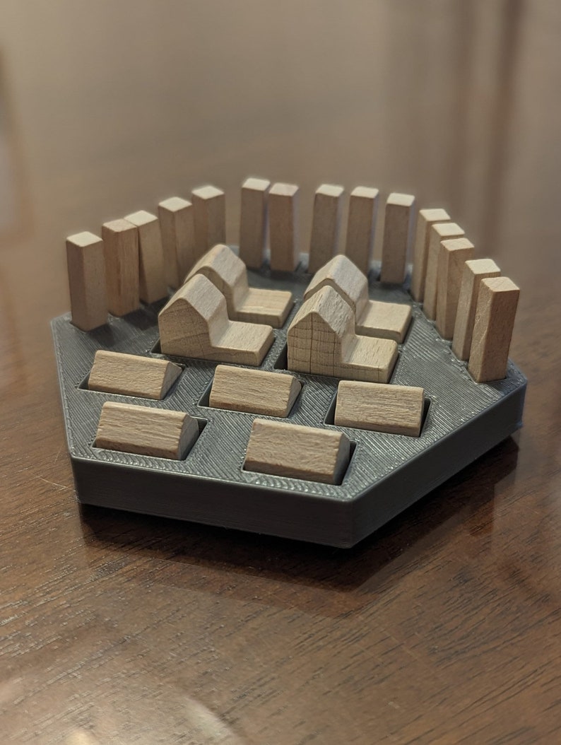 May include: A wooden game board with a hexagonal base and a gray surface. The board has slots for wooden pieces, including houses and fences. The pieces are arranged in a pattern, creating a small village.