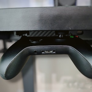 Może przedstawiać: Czarny kontroler do gier wideo jest zamontowany pod biurkiem. Kontroler ma czarny i zielony przycisk na górze i port USB na dole.