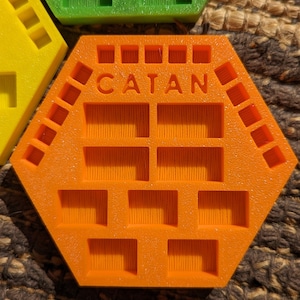 May include: Orange hexagonal game tile with the word "CATAN" printed on it. The tile has multiple square indentations.