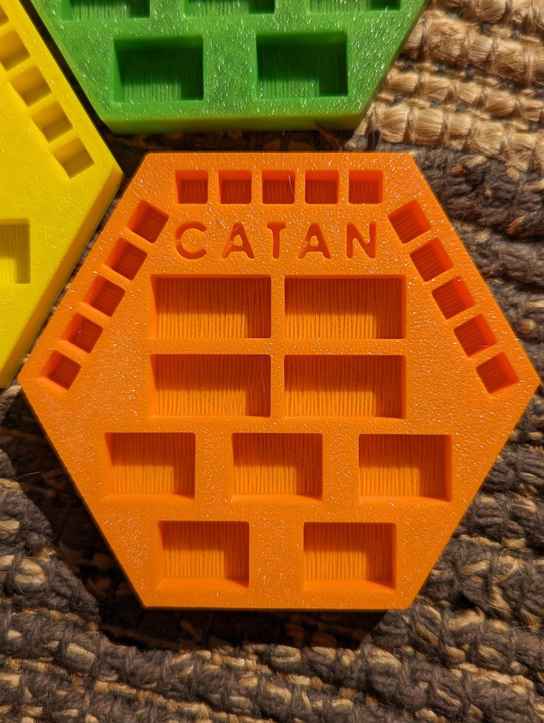 May include: Orange hexagonal game tile with the word "CATAN" printed on it. The tile has multiple square indentations.