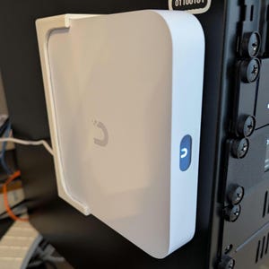 Puede incluir: Un dispositivo electrónico rectangular blanco con un logotipo curvo y un botón azul. El dispositivo está montado en una superficie negra, con tornillos visibles en el lado derecho. El dispositivo tiene un diseño limpio y moderno.