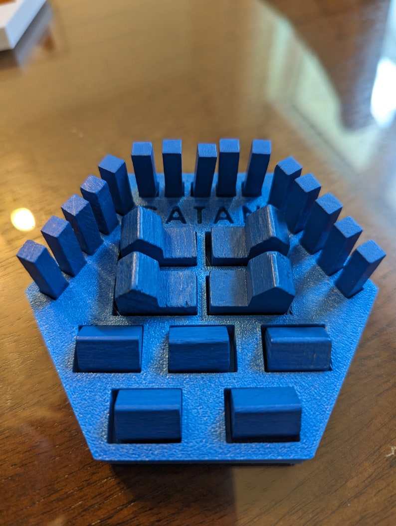 May include: A blue Catan game piece with the word "CATAN" printed on it. The piece has a hexagonal base with a raised center and a row of small blue blocks around the perimeter.