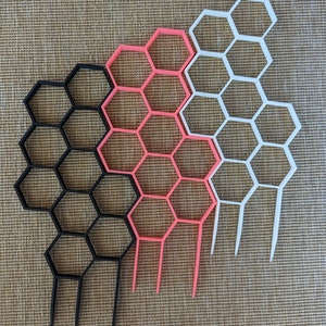 May include: Three honeycomb-shaped wall organisers in black, pink, and white. Each organiser has six rows of hexagonal compartments.