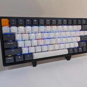 Puede incluir: Teclado mecánico en blanco y negro con teclas retroiluminadas. El teclado tiene un marco negro, teclas blancas y negras y una tecla de escape naranja. Las teclas están iluminadas con una gama de colores.