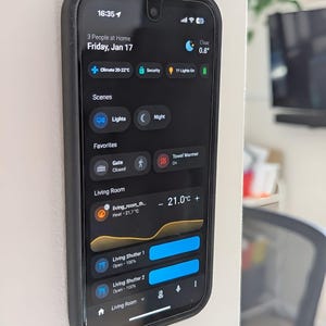 May include: A smartphone in a black case shows a smart home control interface. The screen displays the time, date, weather, and home automation controls for lights, climate, and security. The temperature is displayed as 21.0°C.