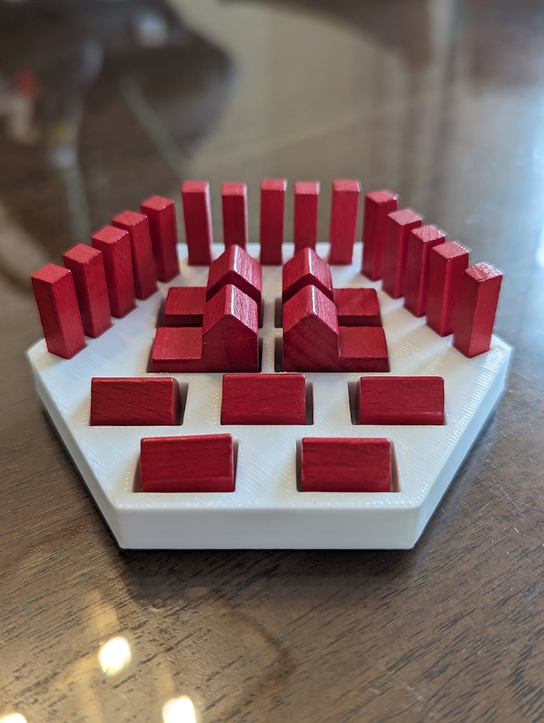 May include: A white hexagonal game board with red wooden blocks arranged in a pattern. The blocks are in the shape of houses and rectangles.