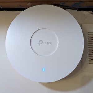 May include: A white TP-Link wireless access point fixed to a wall. The device is circular with a central recessed area and a small blue LED indicator. The TP-Link logo is visible in the centre.