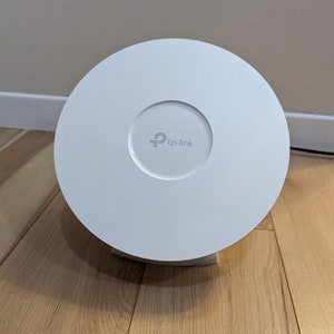 May include: A white, circular TP-Link wireless access point. The device has a central recessed area with the TP-Link logo. It is positioned on a light-coloured wooden surface.