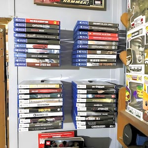 Op de afbeelding: Stapels PlayStation 4-videogame-hoesjes in blauw en zwart. Titels zijn onder meer "Ghostbusters", "Spider-Man" en "Battlefield 1". Een ingelijste Marvel Comics-poster en een PlayStation-tijdschrift zijn ook zichtbaar.