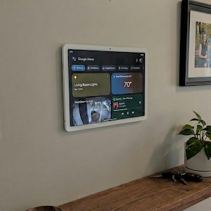 May include: A white Google Home smart home control panel mounted on a wall. The screen displays various home automation controls, including lighting, temperature, and a front door camera feed. The temperature is displayed as 70 degrees.