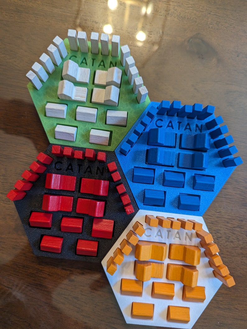 May include: A set of Catan game pieces in different colors, including white, blue, red, and orange. Each piece has the word "CATAN" printed on it.