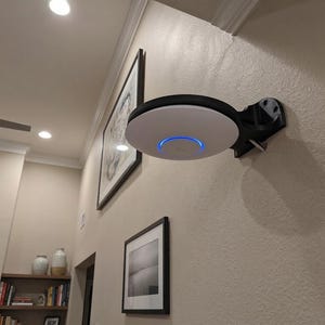 May include: A circular white and black device mounted on a wall, with a blue light. It's attached to a black bracket. The wall is a light beige, with framed pictures and a ceiling fan visible in the background. The device is likely a network access point.