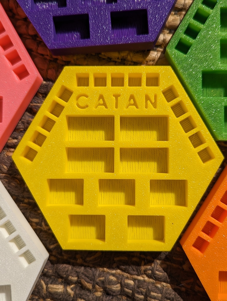 May include: A yellow hexagonal game tile with the word "CATAN" printed on it. The tile has 18 square compartments, each with a textured surface.