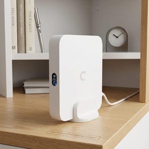 May include: A white Uni-Fi Cloud Gateway stand with a matching white device. The device has a small blue display and a white cable. The stand is on a light wood surface, with the text "UNI-FI CLOUD GATEWAY STAND" visible.