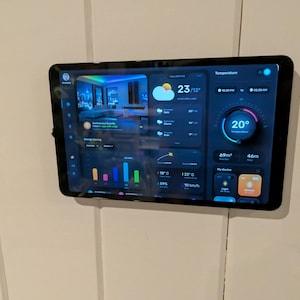 May include: A black tablet mounted on a white wall displays a smart home interface. The screen shows temperature, weather, and energy-saving data. The interface has a dark background with colorful icons and graphs. The temperature is displayed as 20 degrees.