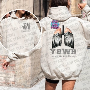 May include: A beige hoodie with a graphic of lungs containing mountain and tree illustrations. The text "YHWH" and "every breath speaks His name" are printed below the lungs. The hoodie also has the text "GRACIE DEAN CREATIONS".