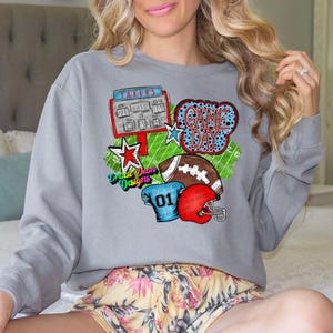 May include: Gray sweatshirt with a colorful football-themed graphic. The design includes a football, helmet, scoreboard, and the words "Game Day." The graphic also features stars and the number "01."