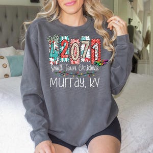 May include: Gray long-sleeve sweatshirt with a colorful graphic design. The design includes the numbers "42071" in a festive style, the text "Small Town Christmas", and "Murray, KY". The sweatshirt has a relaxed fit.