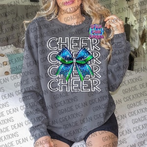 May include: A gray sweatshirt with the words "Cheer" repeated three times in white outlined text. A large green and blue sparkly bow with rhinestones is centered between the words. The words "Gracie Dean Designs" are in a circle on the upper right side of the sweatshirt.