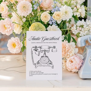 May include: A white sign with black text that reads "Audio Guestbook Leave a message for the newlyweds" with a black and white illustration of a rotary phone. A silver rotary phone sits on a white marble surface next to a bouquet of pastel pink, white, and blue flowers.