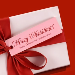 Può includere: Una scatola regalo bianca con un nastro rosso e un'etichetta regalo rosa che dice "Buon Natale" con i nomi Isabella, Charlotte, Eleanor ed Evan.