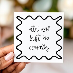 May include: A white square card with a black wavy border and handwritten text that reads "ate and left no crumbs". The card is held by a person with a blurred floral background.