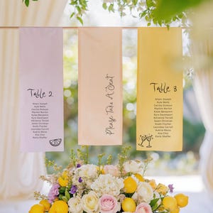 May include: Three table number cards with guest names printed on them. The cards are hung from a gold rod. The card on the left is pink and says "Table 2" with a lemon slice icon. The middle card is peach and says "Please Take A Seat". The card on the right is yellow and says "Table 3" with a champagne flute icon.