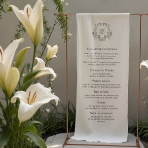 May include: A white fabric banner with a copper stand, listing signature cocktails, sparkling wines, white wines, red wines, beers, and non-alcoholic options. The banner is in front of a white wall with white lilies in the foreground.