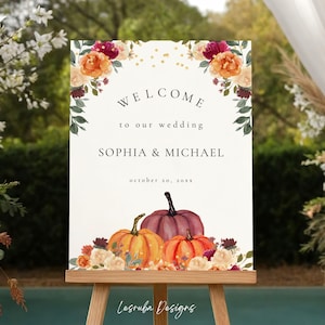 Puede incluir: Un letrero de bienvenida de boda con el texto "WELCOME to our wedding", los nombres "SOPHIA & MICHAEL" y la fecha "october 20, 20xx". El letrero está decorado con arreglos florales y calabazas. Se muestra en un caballete de madera.