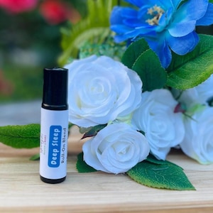 May include: A small, black roll-on bottle with a white label that says "Deep Sleep Roll-On Blend". The bottle is sitting on a wooden surface with white and blue flowers.