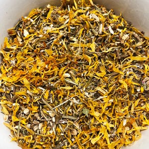 May include: A close-up shot of a white bowl filled with dried herbs and flower petals. The mix features vibrant yellow petals, interspersed with green and brown plant matter, creating a textured, natural appearance. The contents are likely for tea or herbal remedies.