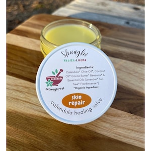 May include: A white jar with a yellow lid labeled "Shuaybi Health & Home" and "skin repair calendula healing salve". The jar contains a yellow salve and the label lists the ingredients: Calendula, Olive Oil, Coconut Oil, Cocoa Butter, Beeswax, Lavender, Tea Tree, and Frankincense.