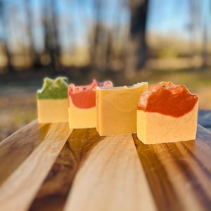 May include: Four bars of handmade soap on a wooden surface. The soaps are green, red, yellow, and orange.