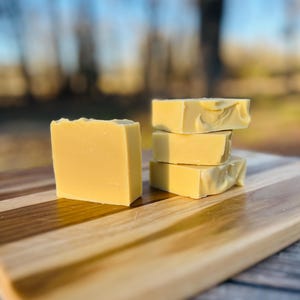 May include: Four bars of handmade soap, stacked and arranged on a wooden cutting board. The soap is a pale yellow color, with a slightly textured surface. The cutting board has a striped pattern of light and dark wood.