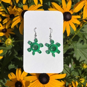 Op de afbeelding: Een paar groene papier-gequilled oorbellen in de vorm van schildpadden. De oorbellen zijn op een witte kaart tegen een achtergrond van gele bloemen.