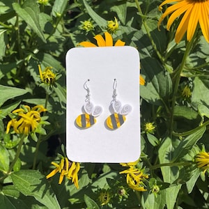May include: A pair of handmade bee earrings. Each earring features a yellow and black striped bee design with silver-colored ear wires and white paper wings. The earrings are displayed on a white card against a backdrop of green foliage and yellow flowers.